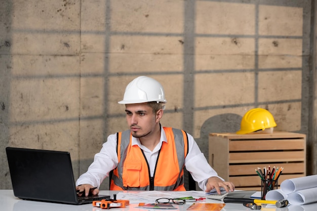 Ingénieur travailleur civil en casque et gilet intelligent jeune beau mec mignon sur ordinateur portable