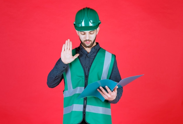 Ingénieur en uniforme vert et casque tenant un dossier bleu, lisant et corrigeant.