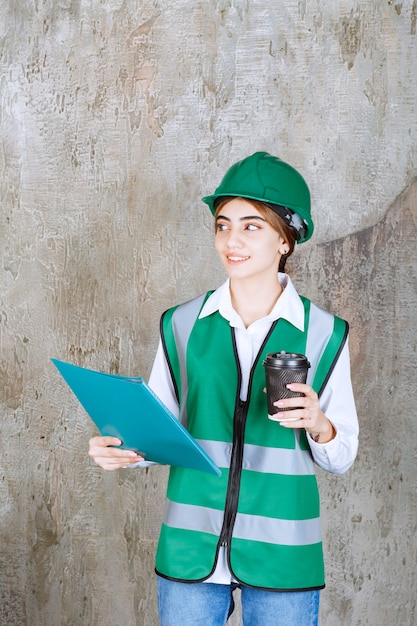 Ingénieur en uniforme vert et casque tenant un dossier de projet vert et une tasse de boisson.