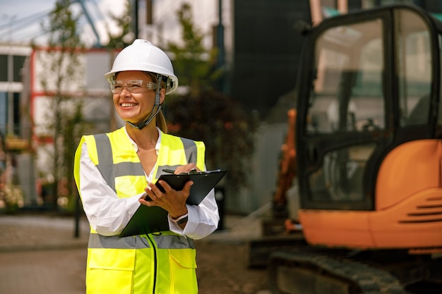 Ingénieure en casque de protection prenant des notes sur le chantier de construction et regardant de côté