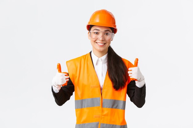 Ingénieure en chef asiatique souriante et satisfaite donnant l'autorisation d'entrer dans un bâtiment ou une entreprise après avoir porté des vêtements de sécurité, des lunettes, des gants et un casque, montrant le pouce levé en signe d'approbation