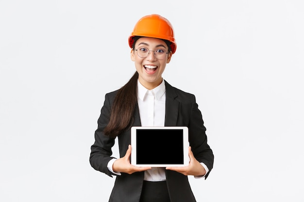 Ingénieure de construction asiatique souriante, excitée et étonnée, architecte en casque de sécurité et costume d'affaires, montrant un écran de tablette numérique avec une expression étonnée, debout sur fond blanc