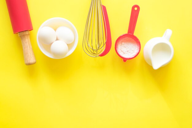 Ingrédients et appareils pour la cuisson. Farine, œufs, lait, rouleau à pâtisserie, fouet sur fond jaune, espace copie, vue de dessus, mise à plat.