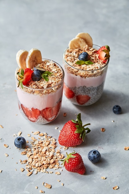 Ingrédients biologiques naturels frais pour cuisiner un petit-déjeuner sain - granola maison, moitiés de fraises, graines de chia, myrtilles et yaourt dans des verres sur une table en pierre grise, place pour le texte.