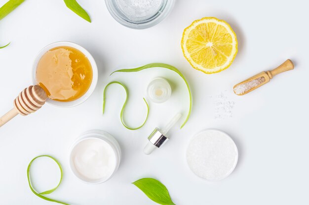 Ingrédients biologiques naturels pour faire des soins de la peau à domicile. Cosmétique nettoyante et nourrissante. Produits de beauté: crème, miel, sel de mer parmi les feuilles vertes sur une surface blanche. Mise à plat, espace de copie pour le texte