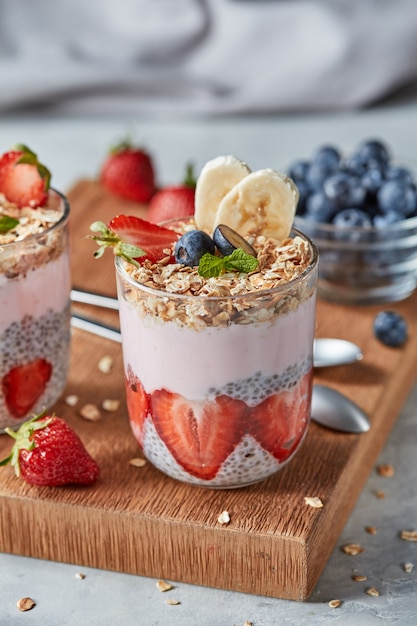 Ingrédients biologiques naturels pour un petit-déjeuner sain dans des verres - granola d'avoine, tranches de banane et de fraise, yaourt aux fruits, graines de chia sur planche de bois, espace de copie.