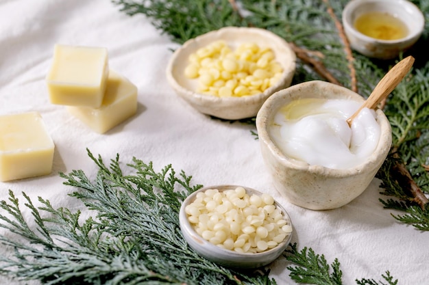 Ingrédients pour la fabrication de cosmétiques naturels faits maison. Crème visage en cours, huiles essentielles, beurre de cacao, cire et savon dans des bols en céramique sur toile de lin blanc avec branches de thuya. Bricolage écologique.