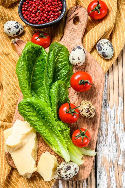 Ingrédients Salade César sur une planche à découper. Laitue romaine, tomates cerises, œufs, parmesan, ail, poivre. Fond blanc. Vue de dessus