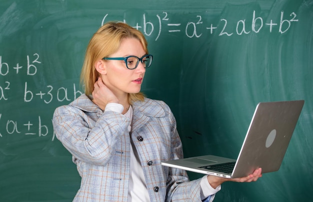 Innovation scolaire Concept d'enseignement à distance Une enseignante porte des lunettes tient un ordinateur portable surfer sur Internet Enseignant femme blonde avec un ordinateur portable moderne sur fond de tableau sur Internet