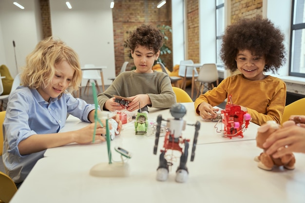 Innovations curieux petits garçons divers souriant tout en étant assis à la table et en vérifiant les jouets robotiques