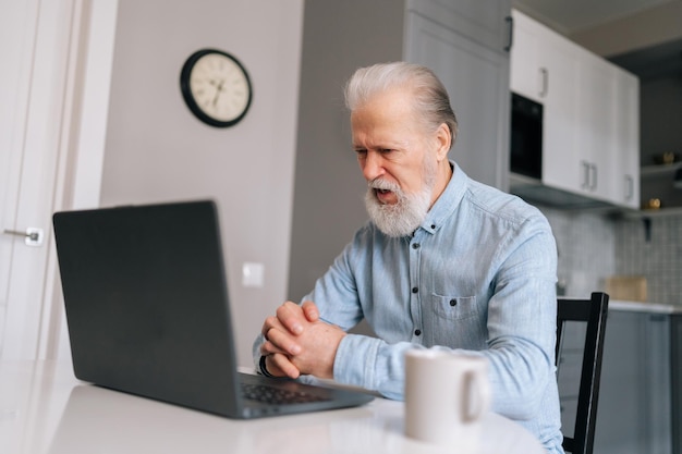 Inquiet en colère barbu mature aux cheveux gris mature homme âgé écoutant un compagnon pendant un appel vidéo sur un ordinateur portable et peut à peine se contenir de jurer assis au bureau avec du café dans la cuisine