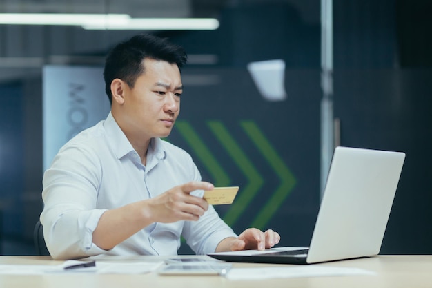 Inquiet jeune homme asiatique assis au bureau dans le bureau, il tient une carte de crédit dans sa main regarde