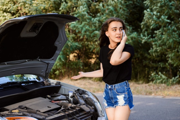 Inquiet jeune jolie fille debout près du capot ouvert de la voiture et téléphonant pour obtenir de l'aide