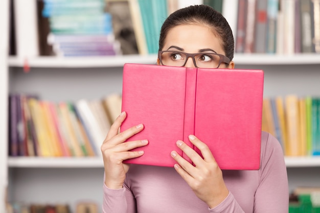 Inquiet pour ses examens. Jeune femme terrifiée regardant hors du livre tout en se tenant devant l'étagère de livre