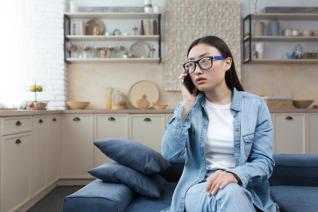 Inquiète et confuse jeune femme asiatique assise sur un canapé à la maison et appelant le service d'urgence sur