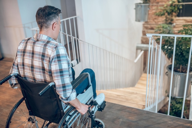 L'insécurité sociale. Vue depuis l'arrière de l'homme en chemise à carreaux sur fauteuil roulant debout dans l'indécision près des escaliers vers le bas à l'intérieur