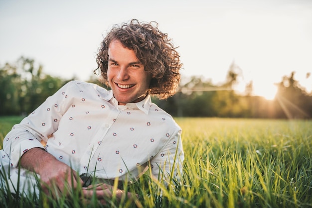 Insouciant heureux beau jeune homme aux cheveux bouclés souriant et riant regardant la caméra assis sur la pelouse verte dans le parc en attendant ses amis Espace de copie pour la publicité Mode de vie des gens