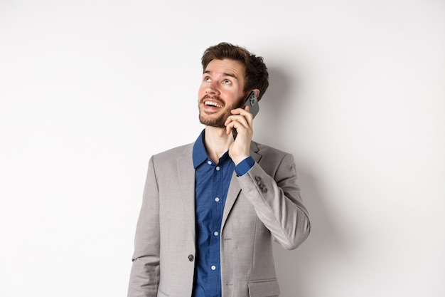 Insouciant jeune homme parlant au téléphone mobile et souriant, levant les yeux avec un visage rêveur, debout en costume sur fond blanc.