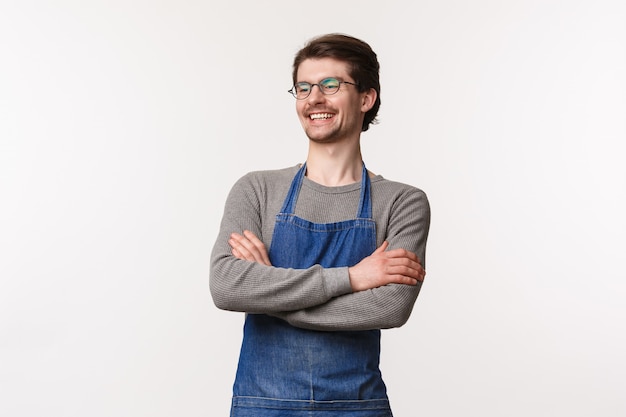 Insouciant souriant jeune barman sortant mâle rire avec un collègue debout en tablier avec les mains croisées sur la poitrine, détournez le regard ravi, gérez votre propre petit café d'entreprise, debout sur un mur blanc