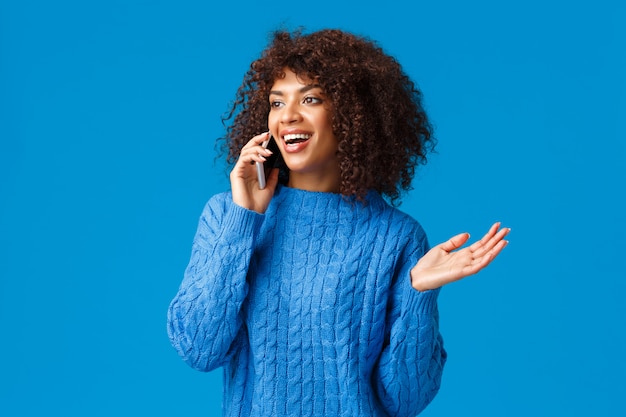 Insouciante bavarde jolie femme afro-américaine en pull d'hiver discuter des vacances récentes et de son voyage à l'étranger station de ski, parler au téléphone, tenant le smartphone près de l'oreille, faire des gestes, sourire