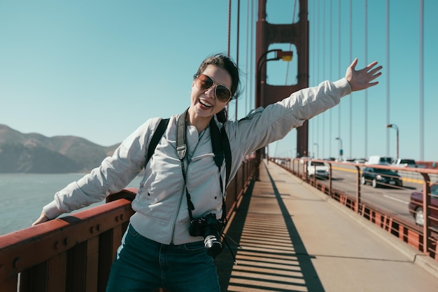 insouciante jeune fille asiatique debout sur le pont de l'état d'or face à la caméra joyeuse rire levant les mains les bras montrant le partage d'une belle vue avec un ciel bleu en été. femme profiter de la nature visites mer.