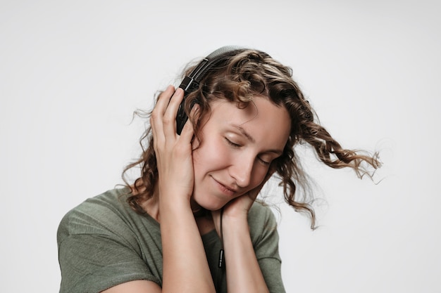 Insouciante joyeuse jeune femme bouclée écouter de la musique préférée avec la main sur ses écouteurs