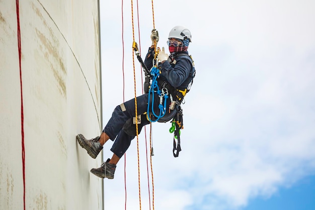 Inspection des travailleurs de sexe masculin portant la première ligne de sécurité de la corde du harnais de sécurité travaillant à un endroit élevé sur le ciel bleu sphérique du toit du réservoir