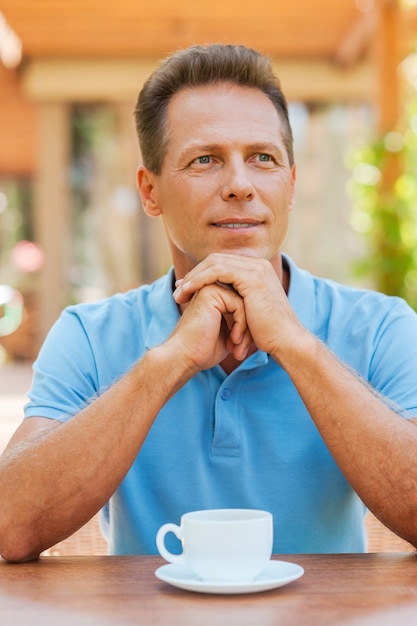 Inspiré d'une tasse de café frais. Homme mûr réfléchi tenant les mains sur le menton alors qu'il était assis dans un café-terrasse avec une tasse de café sur la table