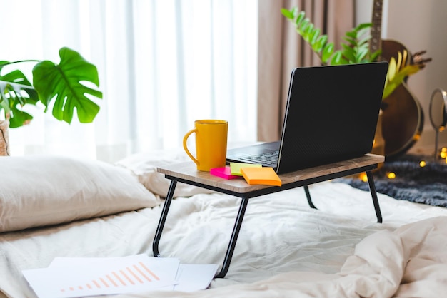 Installation d'un ordinateur portable sur une table portable avec une tasse de café et de thé et des notes autocollantes sur le lit avec une pile de documents et de papiers avec des graphiques et des tableaux placés sur une feuille blanche propre sur le lit dans la chambre