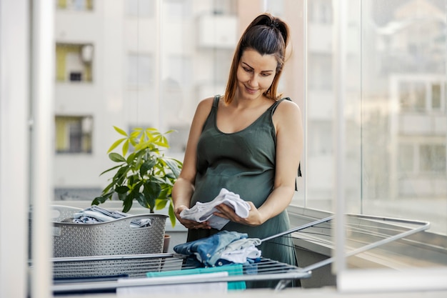 Instinct maternel, tâches ménagères et femmes enceintes. Une future mère excitée prépare des vêtements pour le nouveau-né. Une femme enceinte se tient sur la terrasse et prépare des vêtements pour le nouveau-né