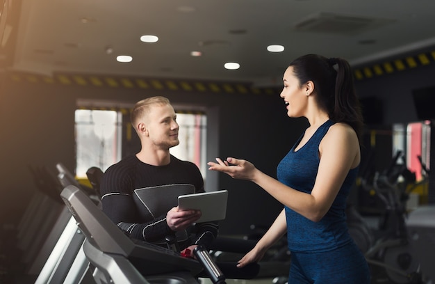 Un instructeur de conditionnement physique aide une jeune femme sur un vélo elliptique. Entraîneur discutant du plan d'entraînement avec une fille sur un tapis roulant. Entraînement cardio en salle de sport, mode de vie sain, espace de copie