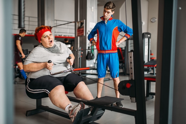 L'instructeur force la grosse femme à travailler sur une machine d'exercice, un entraînement difficile dans une salle de sport.