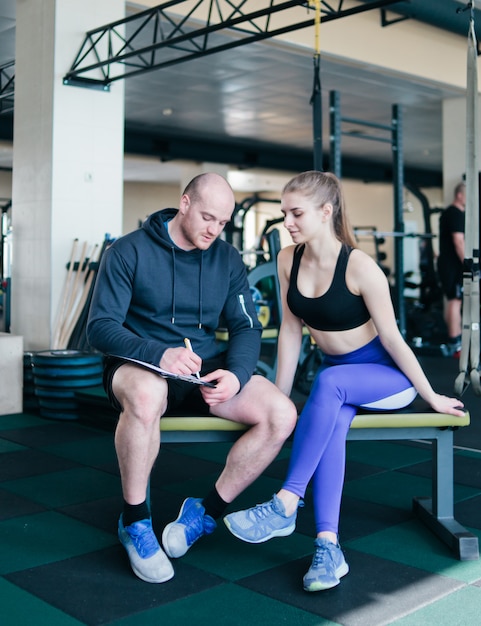 Un instructeur de gym écrit un programme d'entraînement pour une femme blonde sportive assise sur un banc