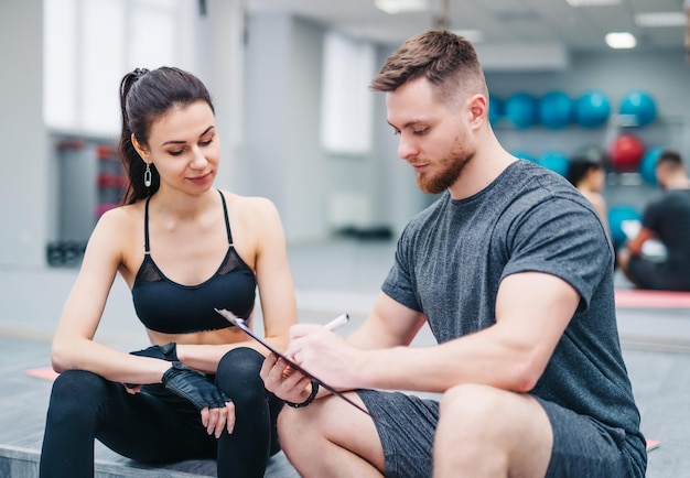 Instructeur masculin musclé écrivant sur un presse-papiers séance d'entraînement spéciale pour une jolie fille sur l'arrière-plan flou