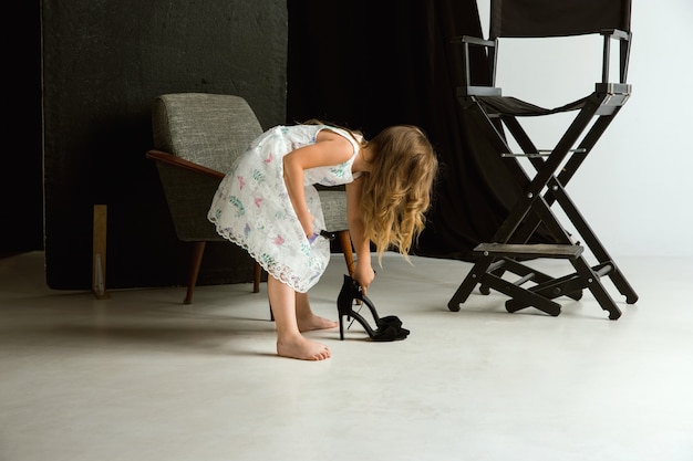 Interaction des enfants avec le monde des adultes. Jolie fille portant les chaussures surdimensionnées de maman et s'habillant pour être plus âgée comme elle l'est. Petit modèle féminin essayant des vêtements à la maison. Enfance, style, concept de rêve.