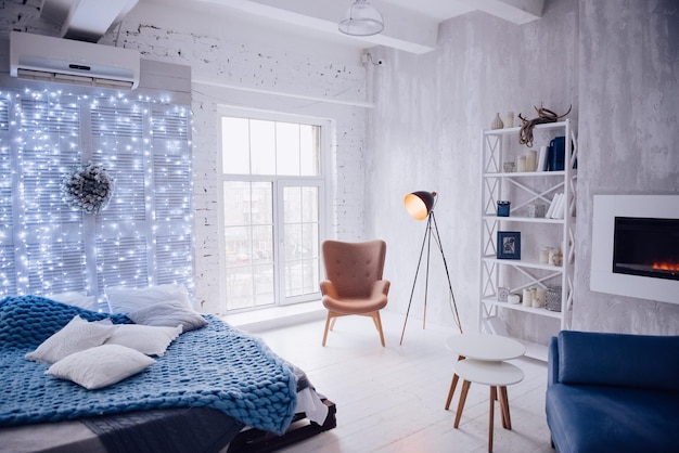 Intérieur du nouvel an de l'appartement. Grandes fenêtres, canapé bleu, cheminée artificielle, lit, lumières, sapin de Noël blanc et bleu, fauteuil et lampadaire élégant. Style loft. Mur en béton. Maison confortable.