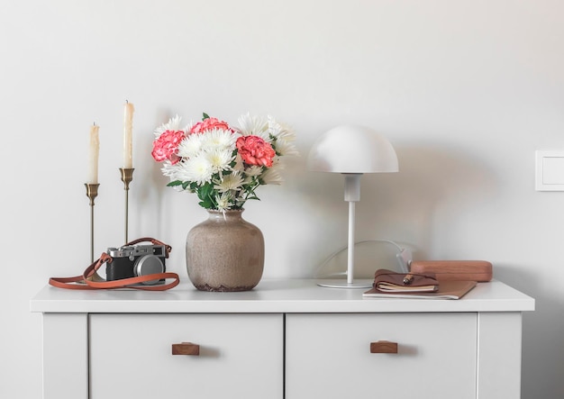 Intérieur du salon bougies fleurs fraîches dans un vase en céramique une lampe de table un cahier et un appareil photo sur une commode blanche dans une pièce lumineuse de style scandinave