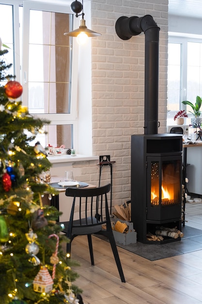 L'intérieur festif de la maison est décoré pour Noël et le Nouvel An dans un style loft avec un poêle noir, un sapin de Noël, un studio chaleureux avec une cuisine blanche brûlant du bois confortable et le chauffage de la maison
