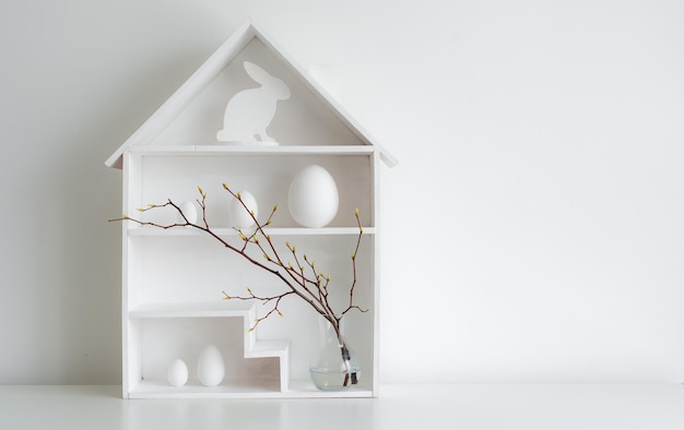 Intérieur minimaliste Springtime Home avec décor de Pâques Branches avec bourgeons en herbe dans un vase en verre sur des étagères en forme de maison Oeufs de Pâques dans un panier de nid sur fond blanc