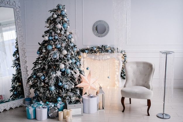 Intérieur De Noël Décoré. Arbre De Noël Avec Des Coffrets Cadeaux Dans Une Salle Blanche. Sapin, Fauteuil, Cheminée Décorée De Guirlandes.