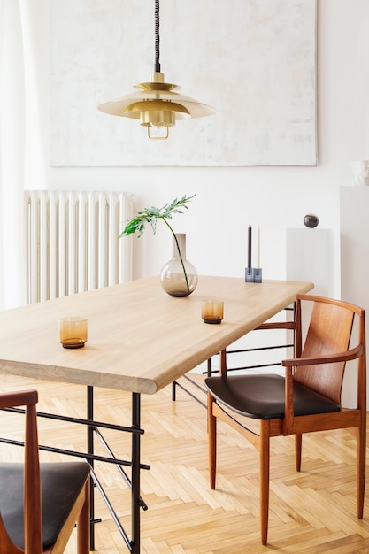 Intérieur de salle à manger élégant et moderne avec table de partage design, chaises, suspension dorée, peintures abstraites et accessoires élégants. Feuilles tropicales dans un vase. Décoration d'intérieur éclectique.