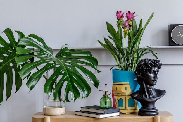 Intérieur de salon minimaliste avec console en bois, belles plantes en pot hipster, feuille, livre, horloge, étagère, décoration, mur gris et accessoires personnels dans une décoration élégante.