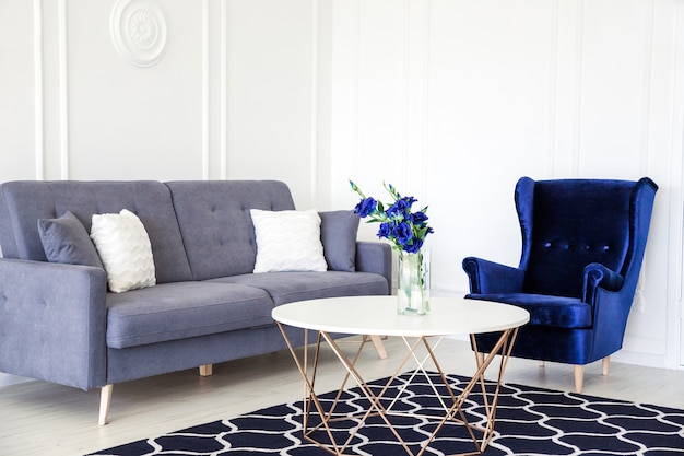 Intérieur de salon moderne - fauteuil en velours côtelé bleu marine, canapé avec coussins, table ronde et vase avec un bouquet de fleurs bleues.