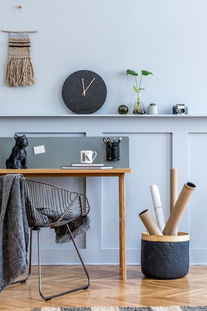 Intérieur de salon scandinave élégant avec bureau en bois, chaise, panneau en bois avec étagère, lampe de table, plantes, horloge noire, fournitures de bureau et accessoires élégants dans un décor de maison moderne.