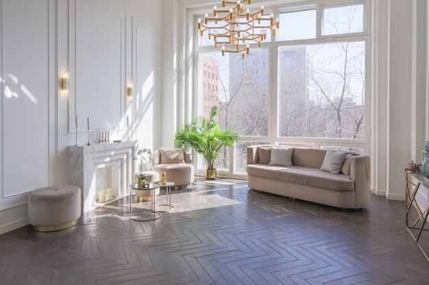 Intérieur très clair et lumineux d'un salon luxueux et confortable avec un mobilier beige doux et chic avec des éléments métalliques dorés, une immense fenêtre au sol et un parquet en bois