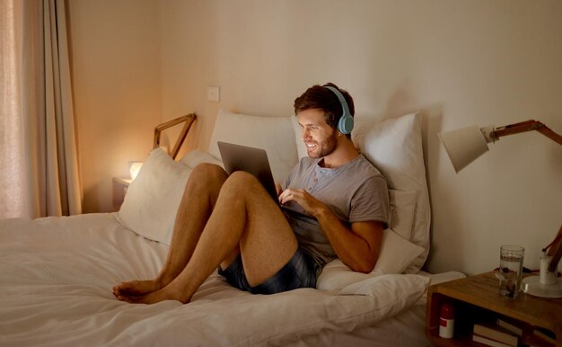 Internet sur ordinateur portable et détendez-vous avec un jeune homme diffusant une vidéo musicale ou un podcast en ligne avec la technologie et assis sur son lit dans la chambre à la maison Profitant de la liberté de la technologie et de la radio pendant le temps libre