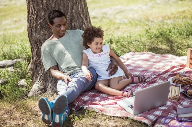 Sur Internet. Petite fille aux cheveux bouclés assise sous l'arbre avec son père et regardant quelque chose en ligne