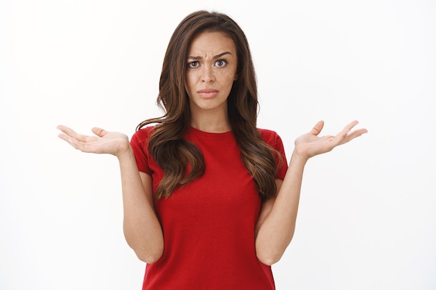 Interrogée et frustrée, une femme brune déçue en t-shirt rouge lève un sourcil sceptique et méfiant, haussant les mains en l'air, grimaçant mécontent, voir une étrange situation absurde