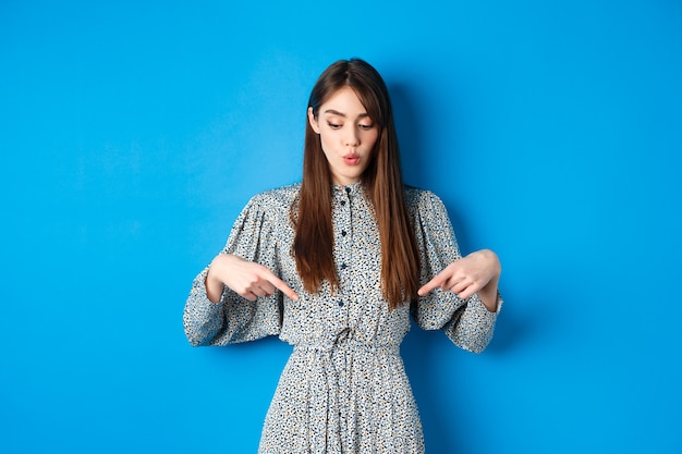 Intrigué belle fille en robe dire wow, regardant et pointant vers le bas à la publicité, montrant la promo avec le visage intéressé, bleu.