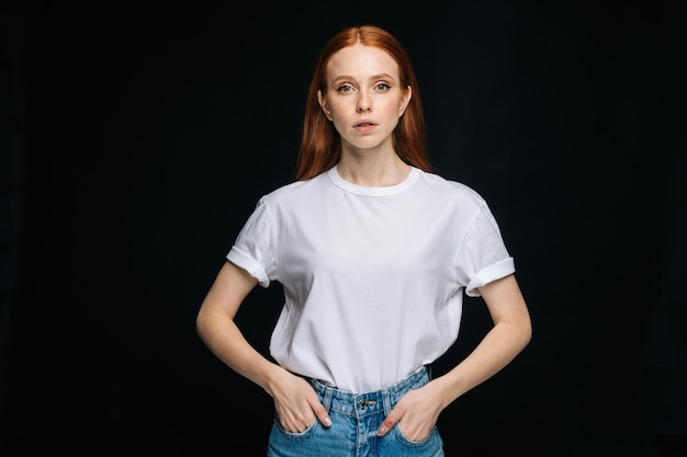 Intshirt de jeune femme sérieuse et pantalon en jean posant avec les mains dans les poches sur fond isolé